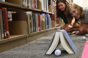 Two students watch a golf ball roll under a book in the library