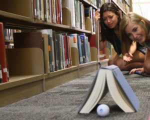 Two students watch a golf ball roll under a book in the library