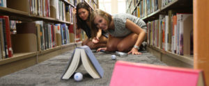 Two students watch a golf ball roll under a book in the library