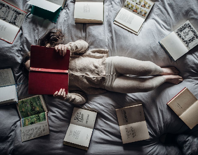 figure lies flat on a bed holding a book up to her face, surrounded by books in the shape of a circle
