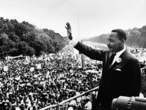 black and white image of Martin Luther King Jr waves to a crowd assembled on the National Mall in Washington, D.C.